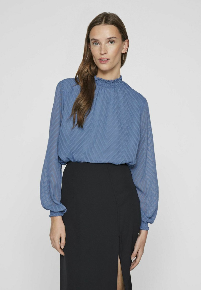 Niebieska zwiewna bluzka z wzorem w zygzak, z wysokim dekoltem i bufkami, zestawiona z czarną spódnicą z bocznym rozcięciem.