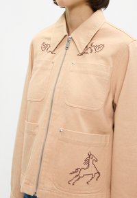 Ανοικτό ροδακινί μπουφάν με φερμουάρ από βαμβάκι που διαθέτει δύο μπροστινές τσέπες. Διαθέτει κέντημα σε σχήμα αλόγου σε μπορντό στην καρδιά και στην τσέπη.