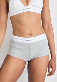 Sivine rebraste hlače Calvin Klein s belim elastičnim pasom, ki vsebuje logotip blagovne znamke in dve vodoravni črti. Mehak tekstilni material.