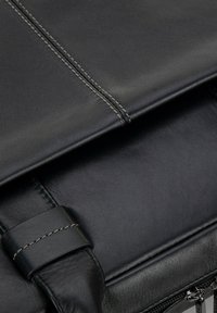 Close-up van de hoek van een zwarte leren tas met gedetailleerde stiksels, een lus voor de schouderband en een gedeeltelijk zichtbare zilveren rits.