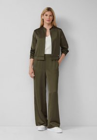Completo sportivo verde oliva con giacca corta e pantaloni larghi, realizzato in un tessuto liscio. Sotto una maglietta bianca; abbinato a sneakers bianche.