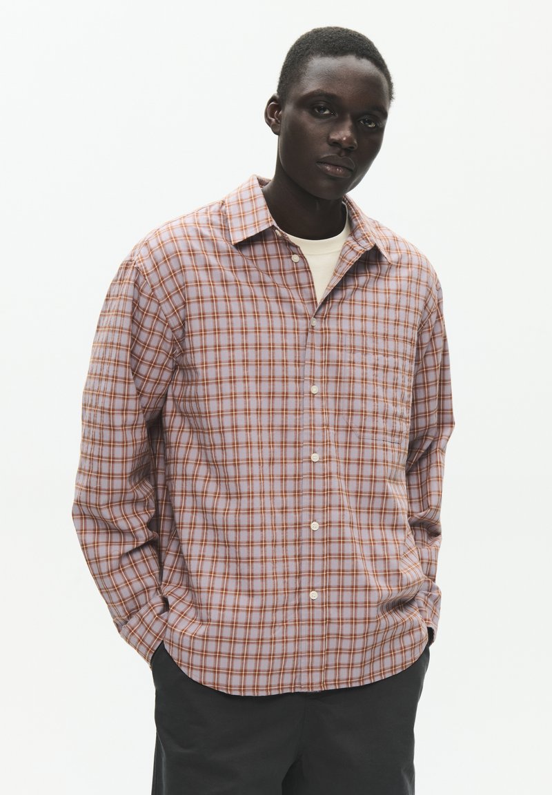 Jeune homme à la peau foncée portant une chemise à carreaux violette claire et orange, debout les mains dans les poches devant un fond blanc uni.