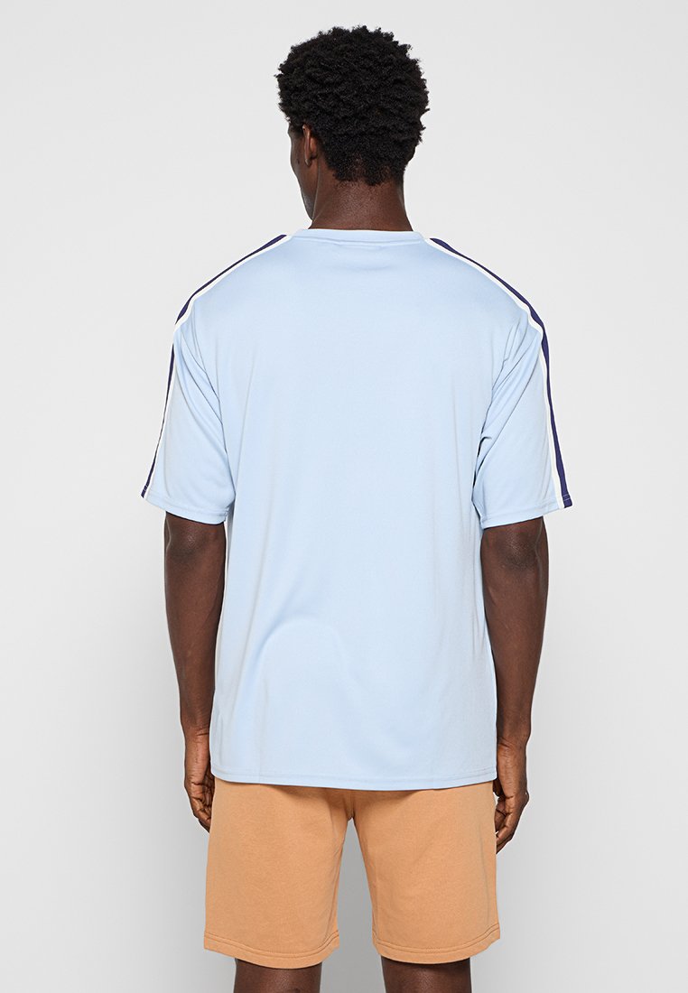 Jasnoniebieska koszulka z krótkim rękawem o luźnym kroju, z granatowymi paskami po bokach i gładką fakturą. Noszona z beżowymi szortami.