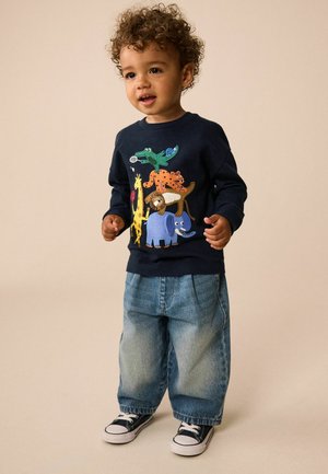 Småbarn med krøllet hår iført en mørkeblå sweatshirt med farverige dyre-stakke, løse blå jeans og sorte sneakers, der står på en beige baggrund.
