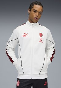 Hvid zip-up jakke med sorte accenter og røde mønstrede detaljer på ærmerne. Har "KING" og fodboldlogoer på brystet.