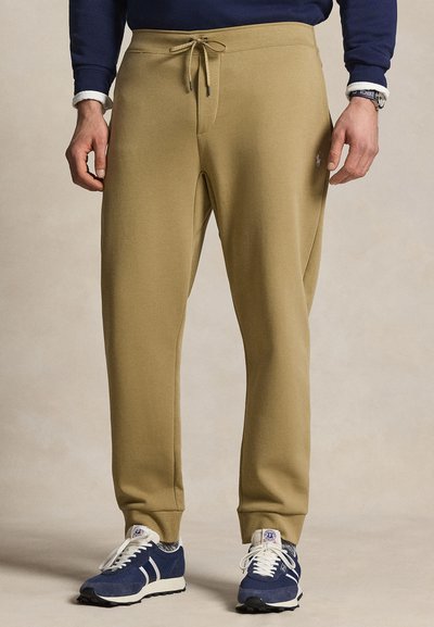 Homme portant un pantalon de jogging beige avec cordon, une chemise à manches longues bleu marine, et des baskets bleu marine et blanches, debout devant un fond neutre.