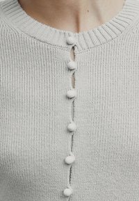 Vaaleanharmaa neulottu paita, jossa on pyöreä kaula-aukko, teksturoitu kangas ja viisi valkoista nappia edessä pienillä nappiaukoilla.