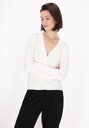 Světle krémový cardigan s V výstřihem, ribbovanou strukturou a pěti knoflíky, kombinovaný s černými kalhotami. Jednoduchý, přiléhavý design.
