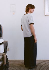 Sivá košeľa s krátkym rukávom a hnedým lemovaním, nosená s voľnými tmavými rifľovými nohavicami. Model stojí pri bielej stene, vedľa vozidla.