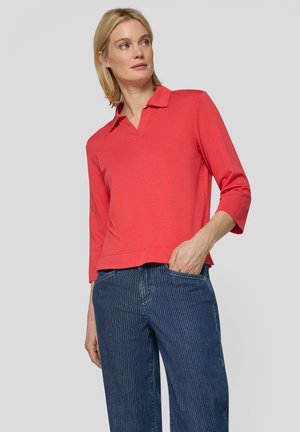 Blonde Frau trägt ein lachsfarbenes Oberteil mit dreiviertel Ärmeln und Kragen sowie eine blaue Jeans mit Nadelstreifen, hat eine Hand in der Tasche und blickt nach links.