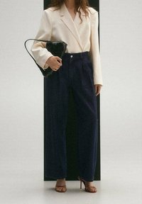 Mujer con un blazer crema, pantalones de pierna ancha de cintura alta en navy, sandalias de tacón en tono beige, sosteniendo un pequeño bolso negro.