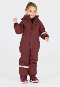 Bordeaux kinderoveralls met hoge kraag, rits aan de voorkant, elastische manchetten en reflecterende strepen. Gecombineerd met donkere sneakers.