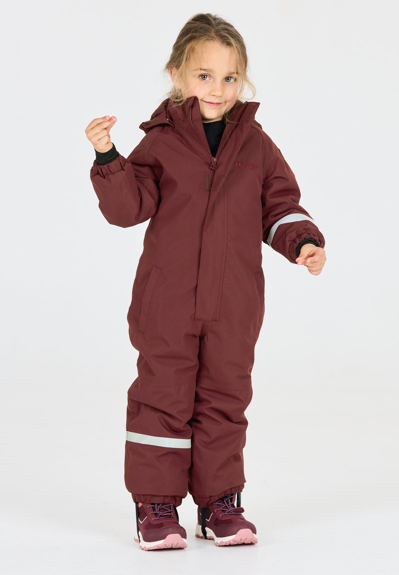 Bordeaux kinderoveralls met hoge kraag, rits aan de voorkant, elastische manchetten en reflecterende strepen. Gecombineerd met donkere sneakers.