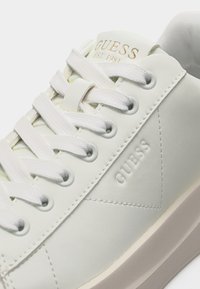 Baskets blanches Guess avec lacets blancs, logo embossé sur le côté et marquage doré "GUESS EST. 1981" sur la languette.