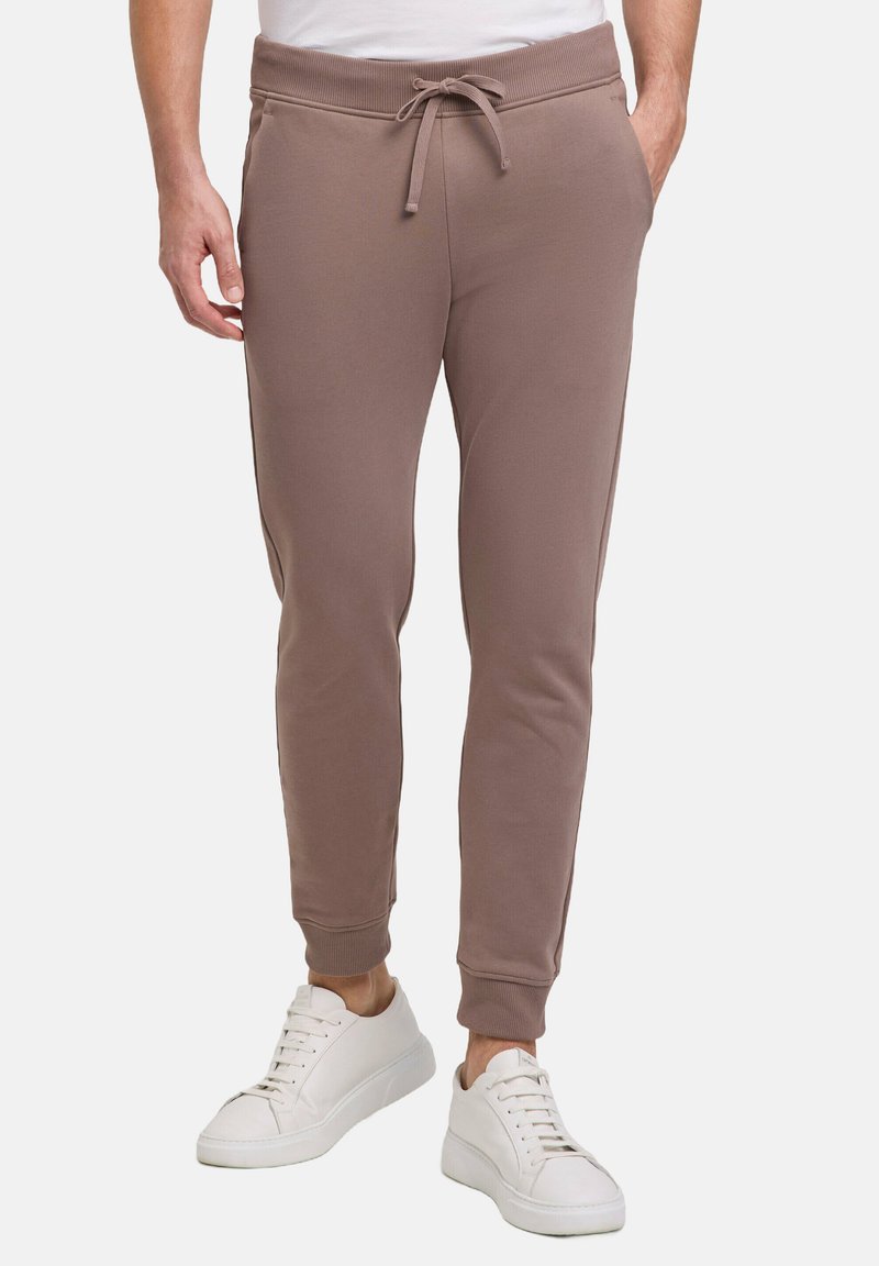 Braune Sweatpants mit Kordelzugbund, gerippten Bündchen und glattem Stoff, kombiniert mit weißen Sneakern.