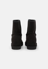 Botas de tobillo de ante negro con textura suave, forma redondeada, cuello acolchado y suela plana de goma. Detalle de costura sutil visible.