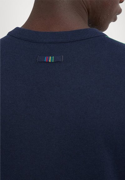 Maglione lavorato a maglia blu navy con una texture a coste, dotato di un piccolo etichetta sul retro con strisce orizzontali rosse, verdi e gialle.