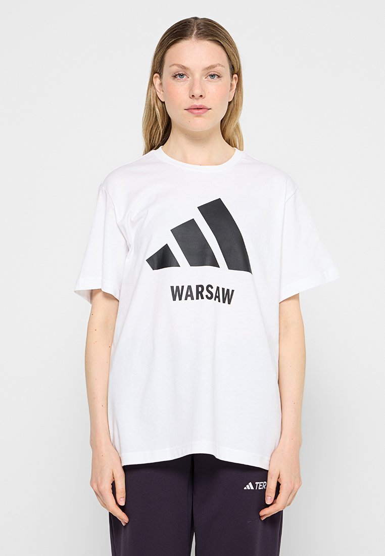 Jeune femme portant un t-shirt blanc taille oversize avec un logo abstrait noir et le texte "WARSAW", associé à un pantalon sombre.