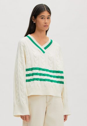 Jonge vrouw met lang zwart haar, gekleed in een off-white kabeltrui met groene strepen en beige broek, staande tegen een witte achtergrond.