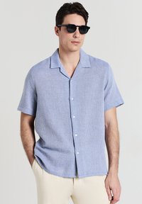Uomo che indossa occhiali da sole neri, una camicia di jeans azzurro chiaro a maniche corte e pantaloni beige, in piedi con una mano in tasca.