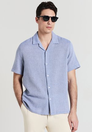 Mann mit schwarzen Sonnenbrillen, hellblauem kurzärmligem Hemd und beigen Hosen, der mit einer Hand in der Tasche steht.