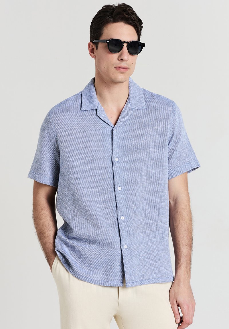 Uomo che indossa occhiali da sole neri, una camicia di jeans azzurro chiaro a maniche corte e pantaloni beige, in piedi con una mano in tasca.