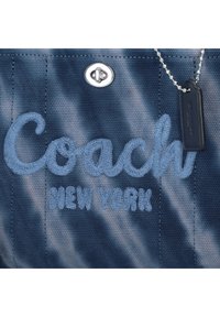 Tissu bleu avec texte brodé "Coach New York" et une fermeture à tourniquet ton argent avec une étiquette en cuir noire attachée sur une chaîne à billes.