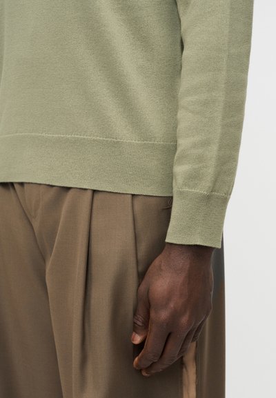 Bras droit et main d'une personne portant un pull vert clair et un pantalon marron ample sur un fond uni.