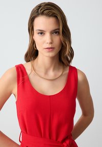 Rotes ärmelloses Kleid mit U-Bootausschnitt und taillierter Taille. Glatter Stoff mit einer subtilen Textur. Das Model trägt eine goldene Creolen-Halskette als Accessoire.