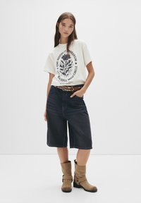 Ung kvinde iført en hvid grafisk t-shirt, mørke denimshorts til knæene, et brunt nittet bælte og lysebrune ruskindsankelstøvler, der står foran en hvid baggrund.