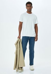 Camiseta de algodón blanca, de manga corta, cuello redondo, combinada con jeans de mezclilla azul. Sosteniendo una chaqueta beige claro. Calzado casual completa el look.