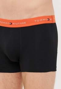 Schwarze Boxershorts mit einem orangen elastischen Bund, der den Text "TOMMY HILFIGER" trägt. Das Material wirkt glatt und hat einen figurbetonten Schnitt.