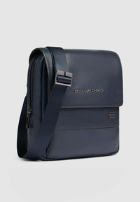 Marineblauwe leren boodschappentas met verstelbare riem, voorklep en subtiel logodetail. Gladde textuur met een ritssluiting compartiment.