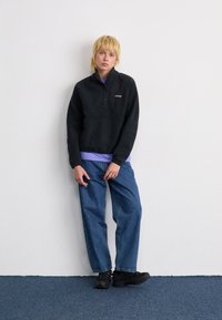 Giovane con capelli biondi, indossa una giacca in pile nera, jeans blu, sneaker nere e una maglietta viola, appoggiato a una parete bianca.