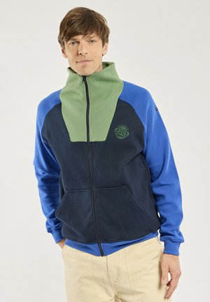 Homme portant une veste polaire à blocs de couleurs vert, marine et bleu, debout les mains dans les poches d’un pantalon beige, sur fond uni.