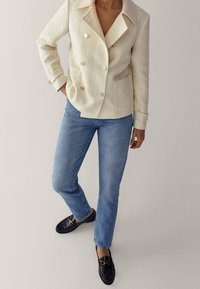 Blazer crema doppiopetto con grandi bottoni dorati, due tasche frontali, abbinato a jeans dritti blu chiaro e mocassini neri.