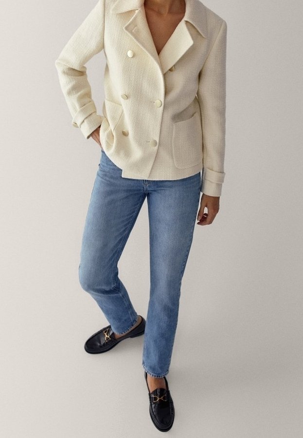 Blazer crema doppiopetto con grandi bottoni dorati, due tasche frontali, abbinato a jeans dritti blu chiaro e mocassini neri.