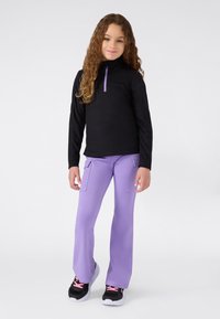 Ragazzina con lunghi capelli ricci, indossa un maglione nero con zip a quarti, pantaloni cargo lavanda e sneaker nere con lacci rosa, in piedi.