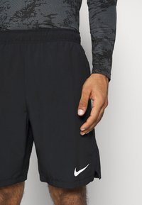 Hombre con pantalones cortos deportivos negros de Nike y una camisa de manga larga con estampado, con la mano relajada al lado, visible el logo de Nike en blanco en los pantalones.