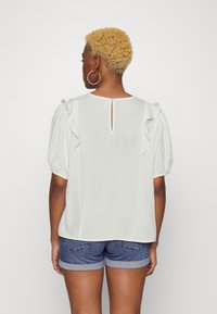 Blusa branca com mangas bufantes e detalhes de babados. Apresenta um fecho nas costas em forma de olho de fechadura. Textura suave, usada com calções de ganga.