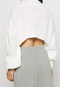 Weißer, verkürzter, gestrickter Pullover mit abgesenkten Schultern, strukturiertem Muster und asymmetrischem Kragen, kombiniert mit grauen, lässig geschnittenen Hosen.