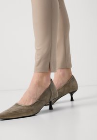 Brune lær kitten-heel sko med spisse tær, med sydd detaljer og en liten hæl, sammen med beige tettsittende bukser.