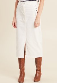 Jupe midi en denim blanc avec des accents de boutons sur le côté et une fente à l'avant. Assortie à des bottes marron montantes. Texture lisse et coupe ajustée.