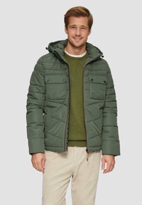 Veste rembourrée verte avec capuche, présentant un design matelassé, deux poches sur la poitrine et une fermeture éclair contrastante. Portée par-dessus un pull vert.