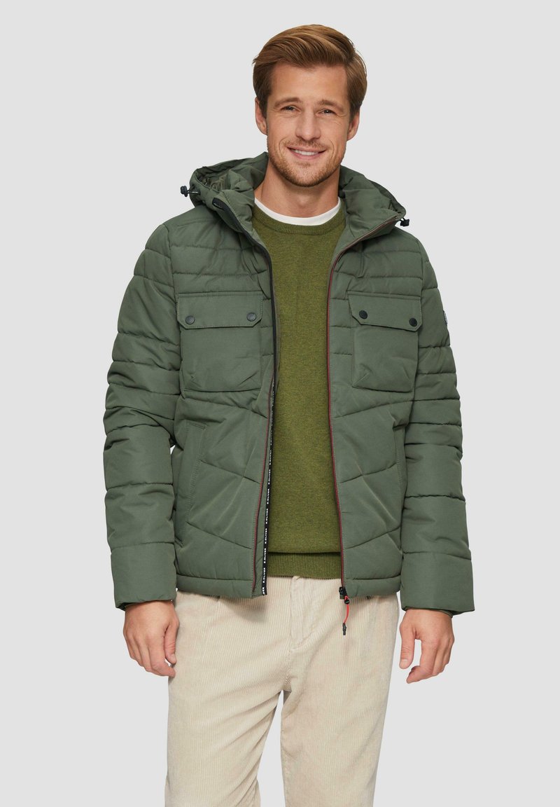 Veste rembourrée verte avec capuche, présentant un design matelassé, deux poches sur la poitrine et une fermeture éclair contrastante. Portée par-dessus un pull vert.