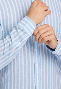 Biała koszula w niebieskie paski z zapięciem na guziki przy mankietach i luźnym kroju. Materiał wydaje się lekki i teksturowany.