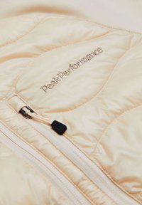 Beige gewatteerde jack met een rits en een zwarte trekker, geborduurd 'Peak Performance' logo in goud op de voorkant, slank ontwerp.