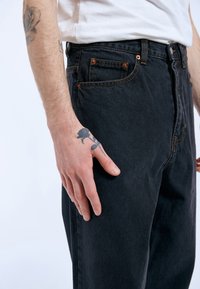 Black denim pants with orange stitching and metal rivets. Features a side pocket and a relaxed fit, shown at hip level with a hand resting on the thigh.