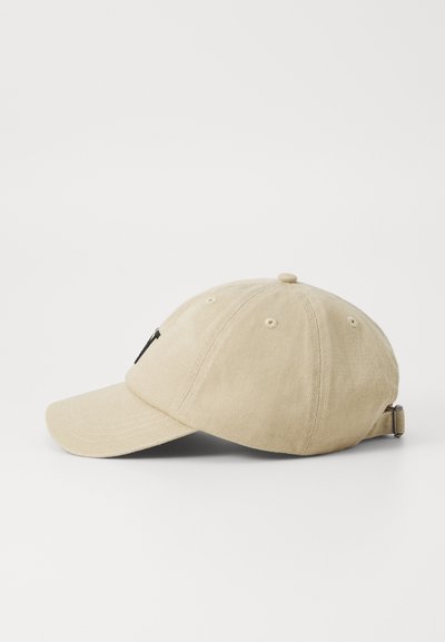 Beige cotton baseball cap with a curved brim, adjustable strap, and a small logo detail on the front. Textured fabric with stitching accents.