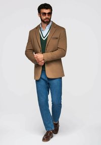 Blazer marrón sobre un chaleco de suéter verde, jeans azules y zapatos marrones. Presenta un pañuelo de bolsillo y gafas de sol redondas.